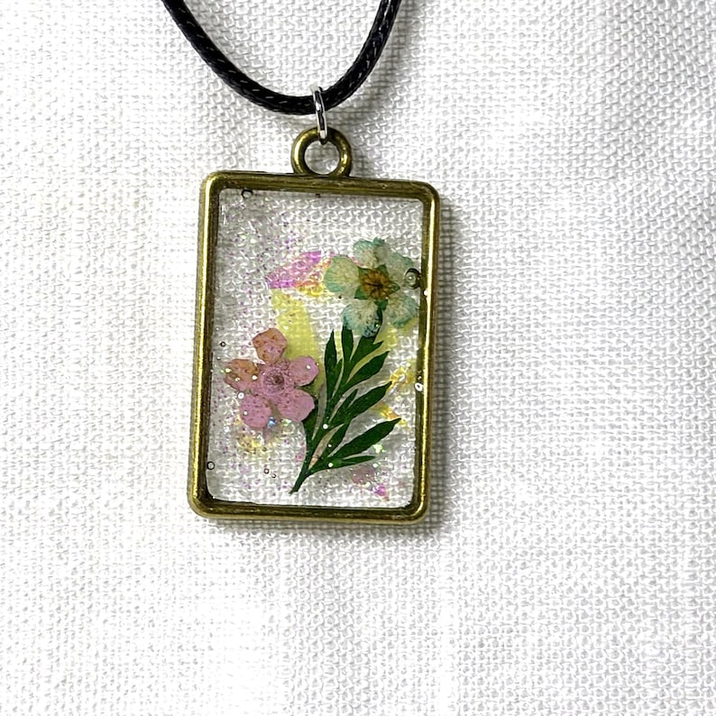 May include: A rectangular pendant necklace featuring a bronze-toned metal frame containing clear resin embedded with two small pressed flowers, one pink and one light blue, along with green leaves and fine iridescent glitter particles.
