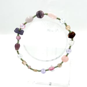 May include: A beaded bracelet featuring a variety of irregularly shaped beads in shades of purple, pink, clear, and brown, interspersed with small metallic green seed beads. The bracelet is arranged in a circular pattern with diverse textures.
