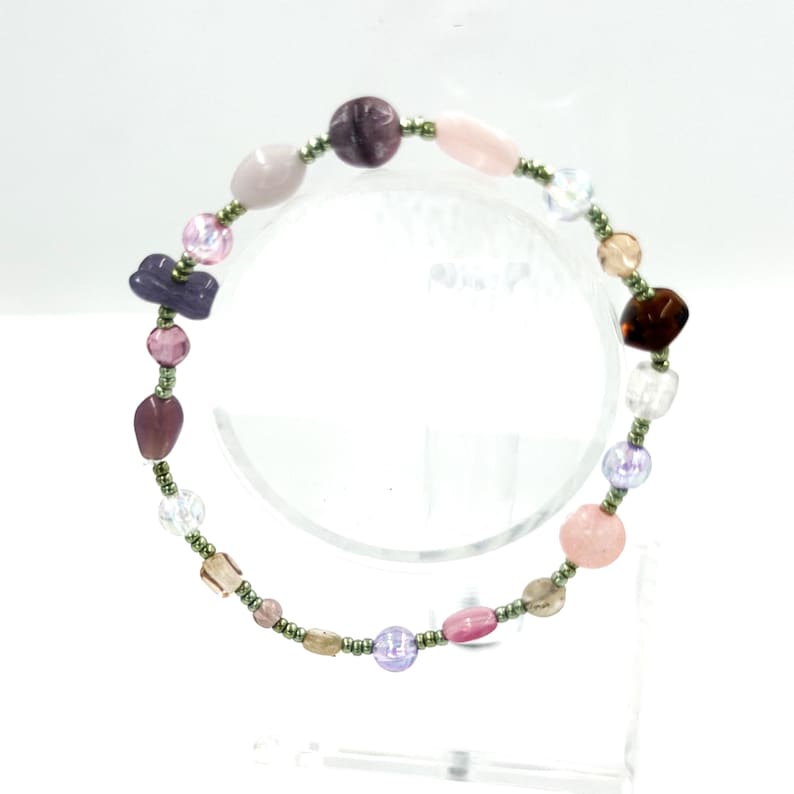 May include: A beaded bracelet featuring a variety of irregularly shaped beads in shades of purple, pink, clear, and brown, interspersed with small metallic green seed beads. The bracelet is arranged in a circular pattern with diverse textures.