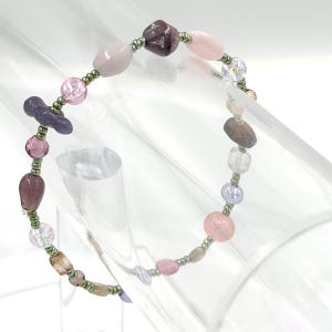 May include: A beaded bracelet featuring a variety of irregularly shaped beads in shades of pink, purple, and clear, interspersed with small metallic green spacer beads. The design includes smooth, faceted, and translucent bead textures.