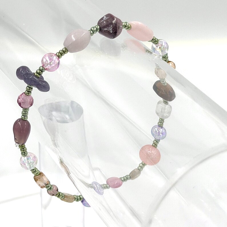May include: A beaded bracelet featuring a variety of irregularly shaped beads in shades of pink, purple, and clear, interspersed with small metallic green spacer beads. The design includes smooth, faceted, and translucent bead textures.