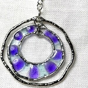 May include: A silver-toned pendant featuring two concentric, irregular circular frames. The inner ring contains translucent, light blue glass segments accented with distinct, rounded purple spots, all held together by a metallic solder frame.