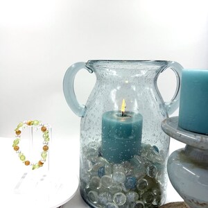 May include: A beaded bracelet with green and brown beads on a clear stand, next to a light blue glass vase with two handles containing a lit teal candle and clear glass pebbles, and a separate teal pillar candle on a gray pedestal base.