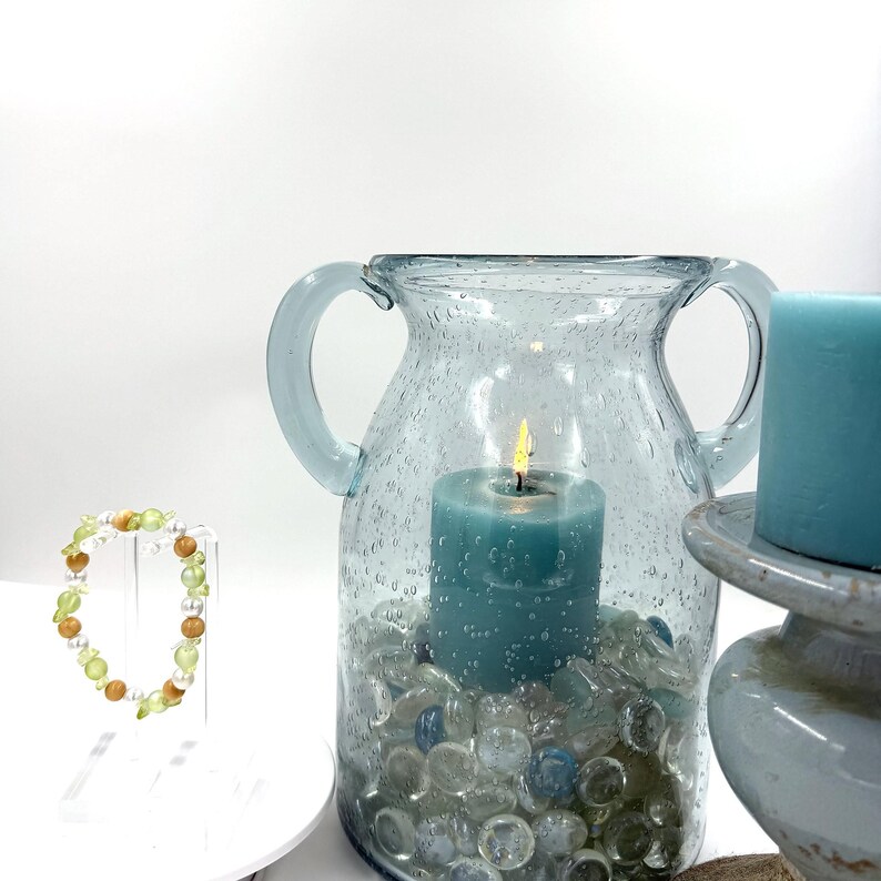 May include: A beaded bracelet with green and brown beads on a clear stand, next to a light blue glass vase with two handles containing a lit teal candle and clear glass pebbles, and a separate teal pillar candle on a gray pedestal base.