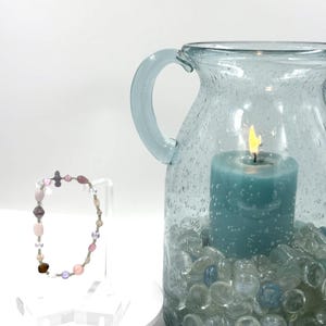 May include: A beaded bracelet featuring a variety of small, multi-colored translucent and opaque beads in shades of pink, purple, brown, and clear, alongside a light blue glass pitcher containing a lit teal candle and clear glass pebbles.