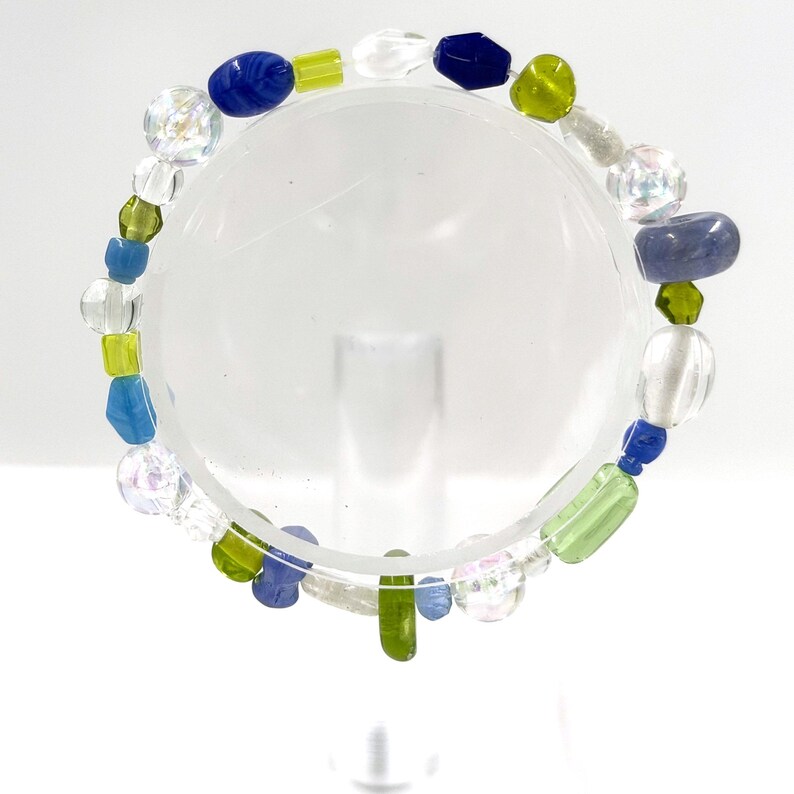 May include: A beaded bracelet featuring a variety of glass beads in shades of blue, lime green, and clear iridescent finishes. The beads are strung on a flexible cord, displaying an assortment of shapes including round, oval, and faceted styles.