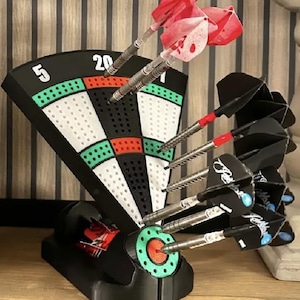 May include: A black dart stand shaped like a dartboard segment with white, green, and red sections labelled with the number 20 and 5. The stand holds several metal darts with red and white flights and black flights featuring white text and blue accents.