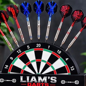May include: A black dartboard display stand with a semi-circular board featuring numbers 5, 20, 1, 18, 4, 13, 14, 9, and 12. The base is labelled LIAM'S DARTS and holds nine darts with red and blue flights and metallic barrels arranged in a fan shape.