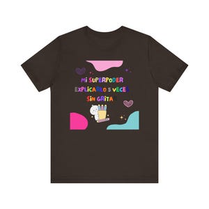 Puede incluir: Una camiseta de manga corta color marrón oscuro con un gráfico que incluye el texto Mi SUPERPODER EXPLICARLO 5 VECES SIN GRITAR. El diseño presenta un pequeño personaje blanco sosteniendo un portalápices, formas abstractas rosas y azules y corazones.