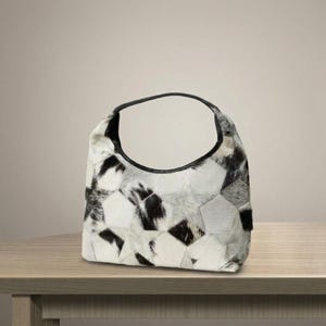 May include: A hobo-style handbag constructed from a patchwork of cowhide panels in shades of white, gray, and black. The bag features a curved top opening with a solid black leather trim along the edge and a single integrated handle.
