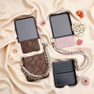 May include: Three foldable phone cases with attached straps. One case has a brown snakeskin texture, one has a pink textured finish, and one is solid black. Each case features a decorative chain strap with white faux pearls.