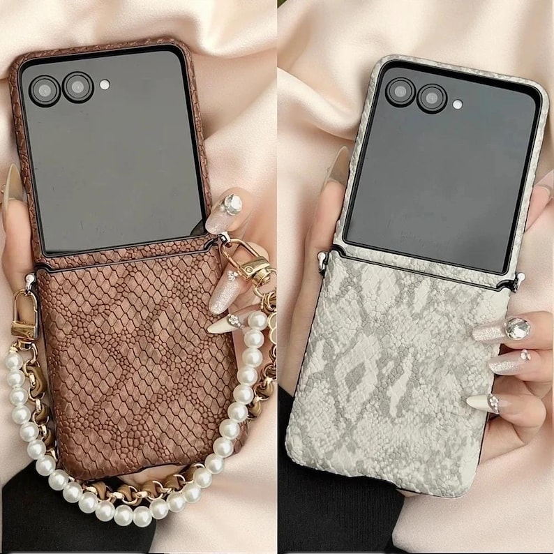 May include: Two foldable smartphone cases with a textured snakeskin pattern. One case is brown and the other is off-white. Both feature a black camera module at the top and a detachable wrist strap made of white faux pearls and gold-tone metal links.