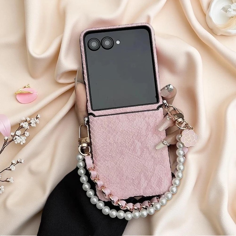 May include: A light pink textured foldable phone case with a dual-lens camera cutout. The case features a decorative wrist strap made of white faux pearls and a braided pink cord, accented with a small pink flower charm and gold hardware.