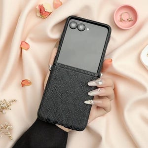 May include: A black foldable smartphone case featuring a textured snakeskin pattern on the lower half and a smooth dark panel on the upper half, designed with two circular camera cutouts and a small flash opening on the back of the device.
