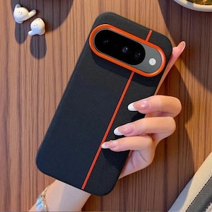 Op de afbeelding: Een zwarte smartphonehoes met een getextureerde afwerking en een glad mat gedeelte. De hoes heeft een opvallende oranje rand rond de cameramodule en een dunne verticale oranje lijn die over de achterkant van de beschermhoes loopt.