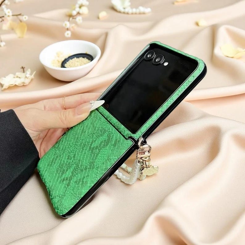 May include: A foldable smartphone case featuring a bright green textured snakeskin pattern on the exterior. The case includes a black frame around the camera lenses and a decorative white pearl beaded wrist strap with a gold-toned metal charm.