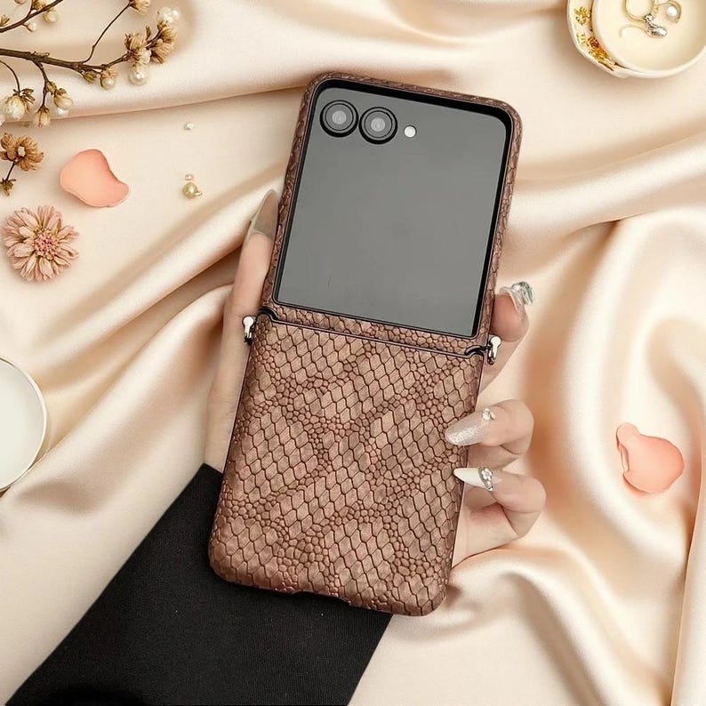 May include: A foldable smartphone case featuring a textured brown snakeskin pattern. The case includes a dark, reflective upper section with two circular camera lenses and a flash, along with integrated metal loops on the sides for attaching a strap.