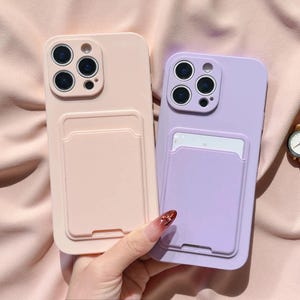 May include: Two smartphone cases, one in a soft peach color and the other in a light lavender shade. Each case features a square camera cutout with three circular lens openings and a rectangular card holder pocket integrated on the back surface.