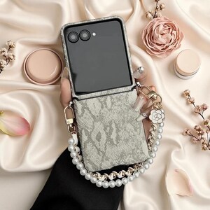 May include: A foldable smartphone case featuring a light grey and white snakeskin pattern. The case includes a detachable wrist strap made of white faux pearls and gold-toned metal links, accented with a small white and gold flower-shaped charm.