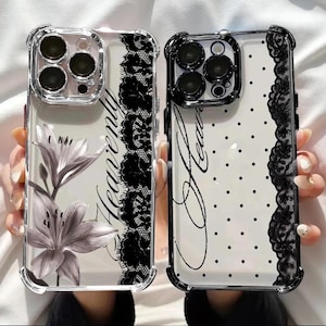 May include: Two clear phone cases with black lace trim. The left case features a gray lily illustration and the word Heavenly in script. The right case has a black polka dot pattern and the word Heavenly in script. Both have black camera bezels.