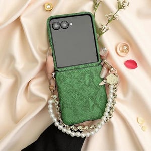 May include: A green foldable smartphone case with a textured snakeskin pattern. The case features a detachable wrist strap made of white faux pearls and gold-toned metal links, accented with a small flower-shaped charm near the attachment point.