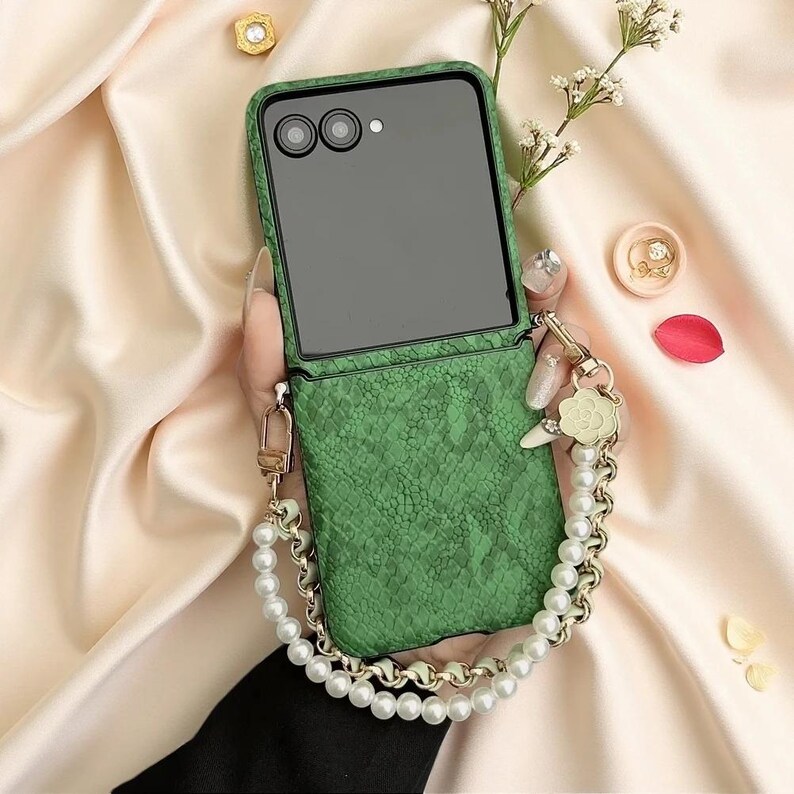 May include: A green foldable smartphone case with a textured snakeskin pattern. The case features a detachable wrist strap made of white faux pearls and gold-toned metal links, accented with a small flower-shaped charm near the attachment point.