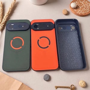 May include: Three smartphone cases are displayed. The first is olive green with an orange circular magnetic ring. The second is bright orange with a black circular magnetic ring. The third is dark gray with a geometric cube pattern on the interior.