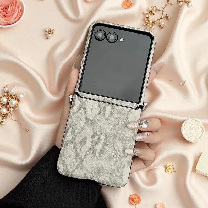 May include: A foldable smartphone case featuring a textured, light grey and white snakeskin pattern. The case includes a black camera module with two circular lenses and a flash, along with integrated metal loops for attaching a carrying strap.