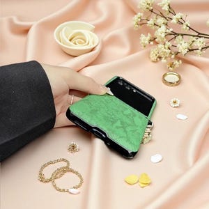 May include: A foldable smartphone case featuring a textured, vibrant green finish on the exterior panels. The case has a black frame and is designed to fit a device that folds in half, with precise cutouts for the camera lenses and ports.