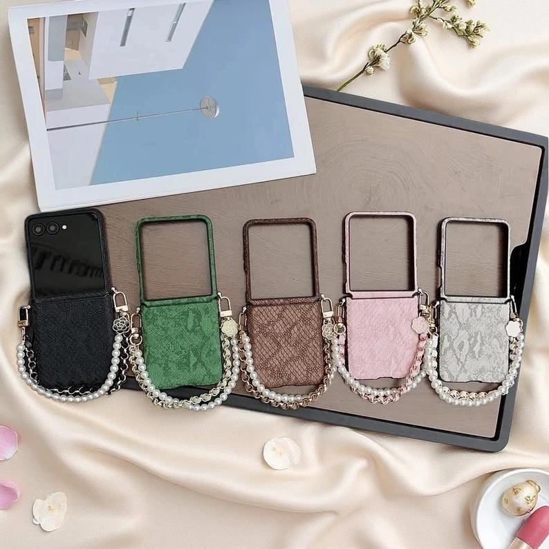 May include: Five foldable phone cases in black, green, brown, pink, and light grey, each featuring a textured snakeskin pattern. Every case includes a decorative gold-tone chain strap adorned with white faux pearls and a small flower charm.