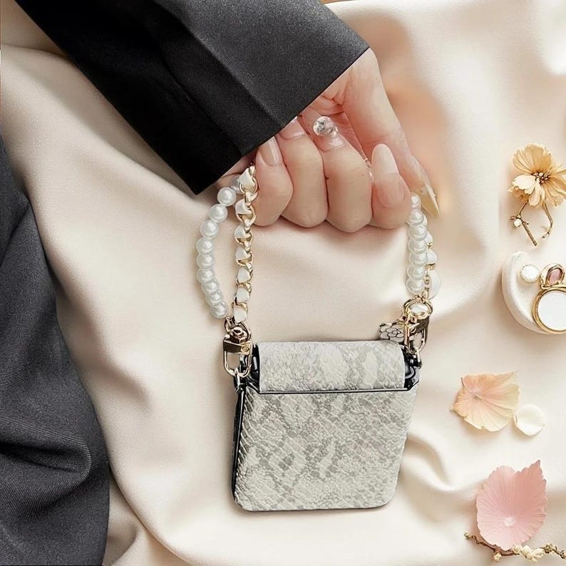 May include: A small, rectangular case featuring a light grey and white snakeskin pattern. The case includes a decorative handle made of white faux pearls and a gold-toned chain, attached with gold-coloured metal clasps on both sides of the top edge.