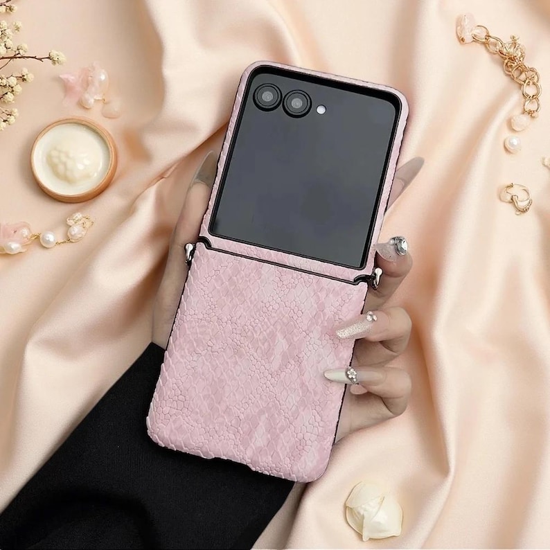 May include: A foldable smartphone case featuring a textured, light pink snakeskin-patterned exterior. The case includes a black frame around the screen area, two circular camera lenses, and small metal loops on the sides for attaching a strap or accessory.