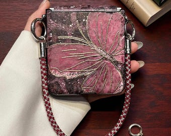 May include: A square, foldable phone case featuring a dark, textured background with a large, embossed pink flower design accented by gold lines. The case includes a detachable strap made of small, alternating red and clear beads with silver hardware.