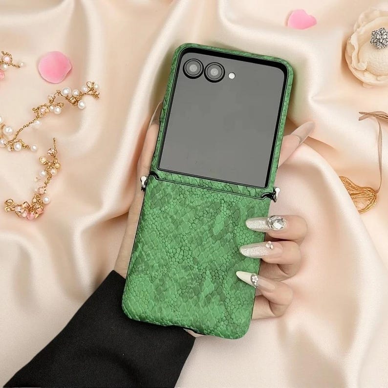 May include: A foldable smartphone case featuring a vibrant green snakeskin texture. The upper portion of the case is black and houses two circular camera lenses and a small flash. The case includes small metal loops for attaching a strap.