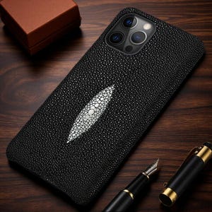 Könnte beinhalten: Eine schwarze Smartphone-Hülle mit texturierter Rochenhaut-Optik und einer markanten, länglichen weißen Einlage in der Mitte. Die Hülle hat eine Aussparung für ein Triple-Kamera-System und liegt auf einer dunklen Holzoberfläche.