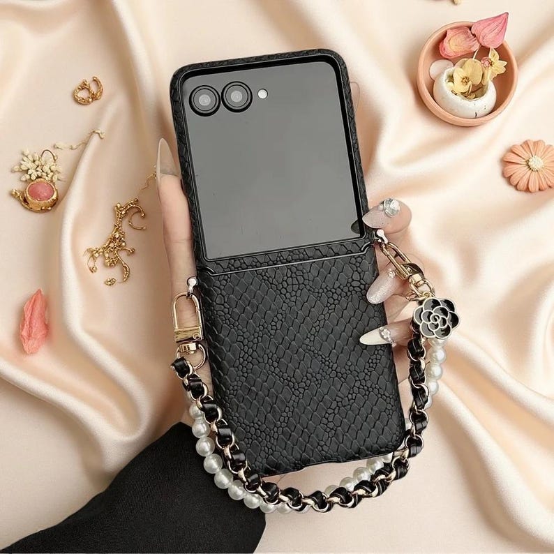 May include: A black foldable smartphone case with a textured snakeskin pattern. The case features a detachable wrist strap made of white faux pearls interwoven with a black leather-like cord, accented by a black and silver flower-shaped charm.