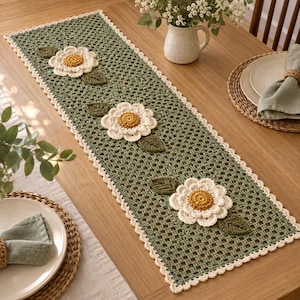 Puede incluir: Un camino de mesa rectangular de ganchillo en color verde salvia con un patrón de rejilla y tres flores blancas grandes y dimensionales con centros amarillos. Cada flor está acentuada por dos hojas de ganchillo verde oscuro. Borde festoneado crema.