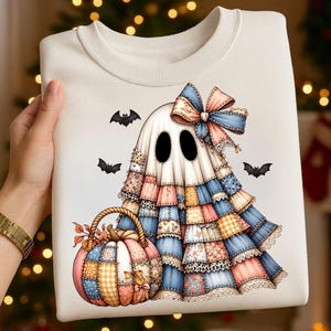 Könnte beinhalten: Ein cremefarbenes Sweatshirt mit einer Grafik eines Geistes, der ein Kleid im Patchwork-Stil mit blauen, rosa und gelben Blumenmustern trägt, dazu eine große Schleife auf dem Kopf und ein passender Patchwork-Kürbiskorb, umgeben von drei kleinen schwarzen Fledermäusen.