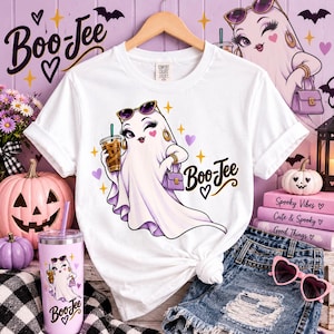 May include: A white short-sleeve t-shirt featuring a graphic of a cartoon ghost wearing sunglasses, holding a coffee cup and a purple handbag, with the text Boo-Jee. The design includes small stars and hearts in purple, yellow, and pink colors.