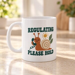 May include: A white ceramic mug featuring a graphic of a brown snail holding a green telephone receiver. Above the snail is a yellow sun and two green leaves. The text "REGULATING" appears in dark green above the snail, and "PLEASE HOLD" below.