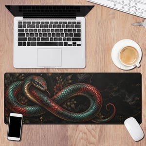 Puede incluir: Una alfombrilla de escritorio rectangular con una serpiente enroscada de escamas iridiscentes rojas y verde azulado sobre un fondo oscuro y sombrío con sutiles elementos florales y geométricos, situada bajo un portátil, un smartphone y una taza.