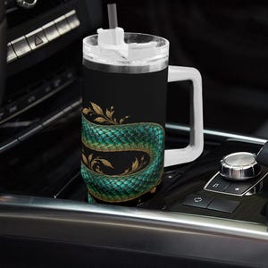 Puede incluir: Una taza de viaje negra con asa blanca y tapa transparente con pajita. La taza muestra un gráfico de una serpiente verde enroscada con escamas detalladas y detalles de hojas doradas envueltas alrededor del cuerpo cilíndrico.