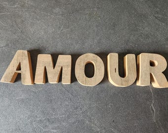 May include: Five individual capital letters made of light-colored wood spelling the word AMOUR. Each letter has a natural grain texture and a matte finish, standing upright to form the complete word in a clean, sans-serif block typeface.