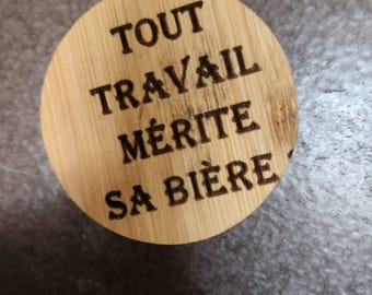 May include: A circular light brown bamboo coaster featuring the laser-engraved black text TOUT TRAVAIL MÉRITE SA BIÈRE. The wood grain is visible across the surface of the round object, which displays a natural texture and a clean, flat design.