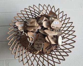 May include: A collection of light-colored wooden keychains in heart and circular shapes, each featuring engraved French text such as 'Meilleur Papy du Monde' and 'Je t'aime Maman', all contained within a star-shaped woven basket with a natural finish.