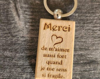 May include: A rectangular light-colored wooden keychain featuring a silver metal ring. The wood is engraved with a heart symbol and the French text: Merci de m'aimer aussi fort quand je me sens si fragile. The surface shows a natural wood grain.