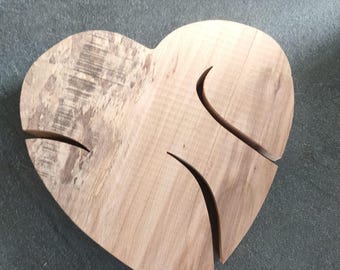 May include: A heart-shaped wooden object with a natural light brown grain and texture. The surface features two distinct curved cutouts that divide the heart into three separate pieces, showcasing the raw wood finish and visible grain patterns.