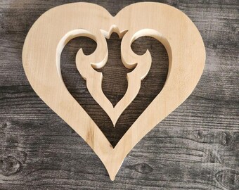 May include: A light-colored, unfinished wooden heart cutout featuring a decorative, symmetrical filigree-style cutout design in the center. The wood grain is visible across the smooth surface of the heart, which has a pointed bottom and rounded top.