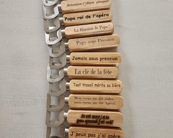 May include: A collection of fourteen bottle openers with light-colored wooden handles. Each handle features unique black text in French, including phrases like Libère la pression, Cheffe sous Pression, and Papa roi de l'apéro, attached to silver metal openers.