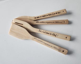 May include: Four light-colored wooden kitchen utensils, including a spatula and spoons, each featuring laser-engraved French text on the handle. The inscriptions read: En cuisine on m'appelle pas Maman, on dit OUI Chef!, La cuillère magique de Maman, Ici c'est moi le Chef!, and La cuillère magique de Maman.